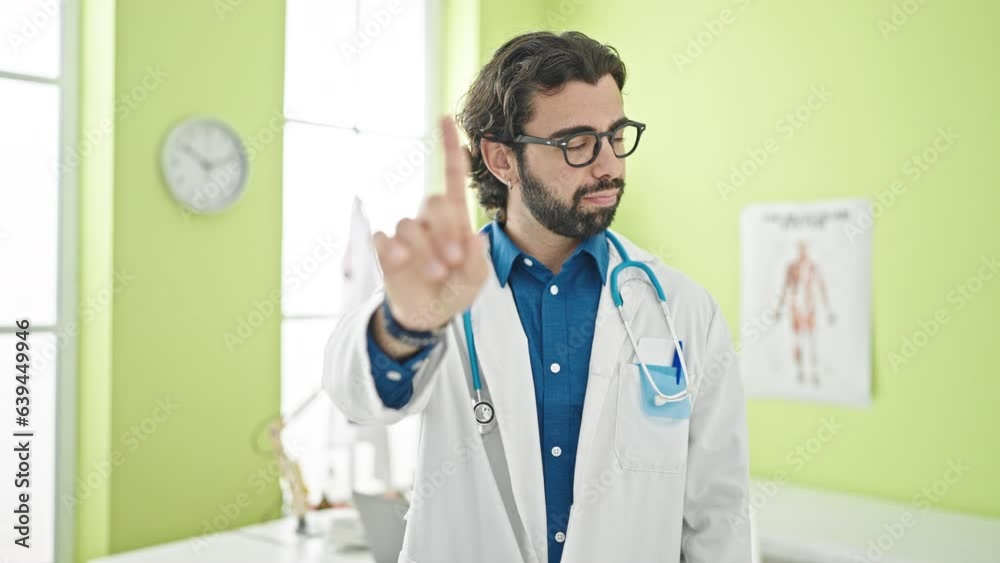 Young hispanic man doctor standing with serious expression saying no with finger at the clinic