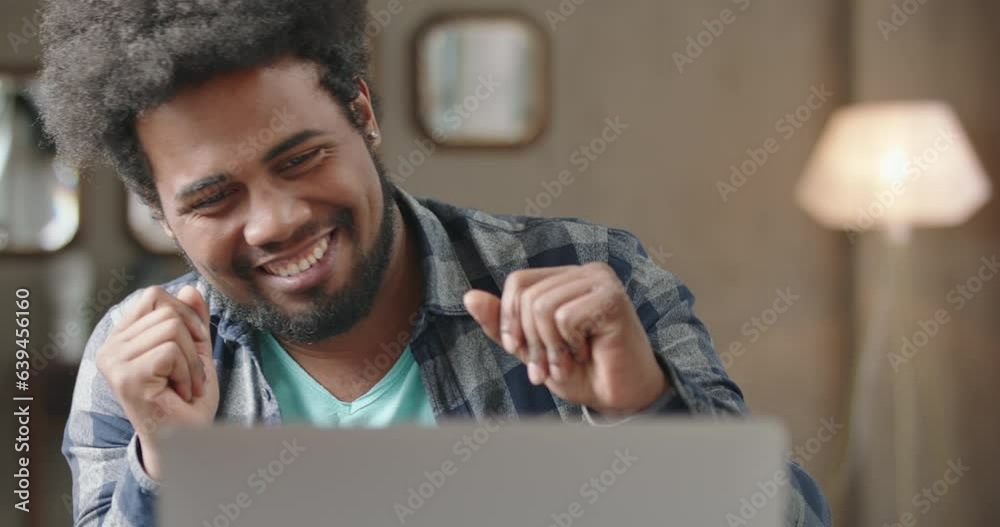 Young black man working on laptop. Man expressively rejoices, laughs, dances merrily in good happy mood. Luck, victory, winning in game. Concept of betting, gambling. Black man wins lottery.