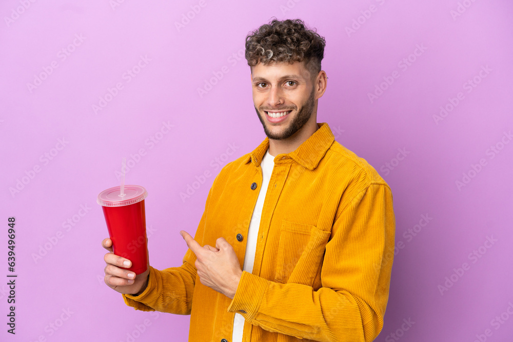 Young blonde man holding soda isolated on purple background pointing back Young blonde man holding soda isolated on purple background pointing back