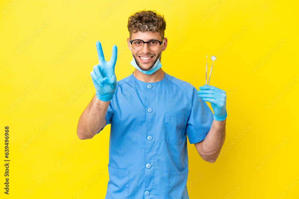 Dentist blonde man holding tools isolated on background smiling and showing victory sign Dentist blonde man holding tools isolated on background smiling and showing victory sign
