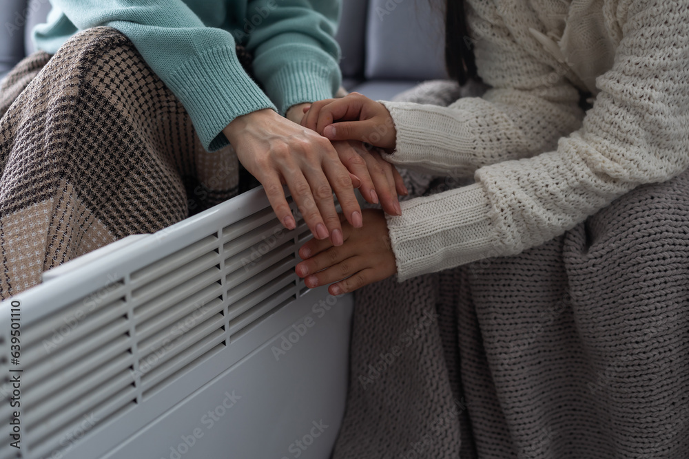 Mother and child warming hands near electric heater at home, closeup.