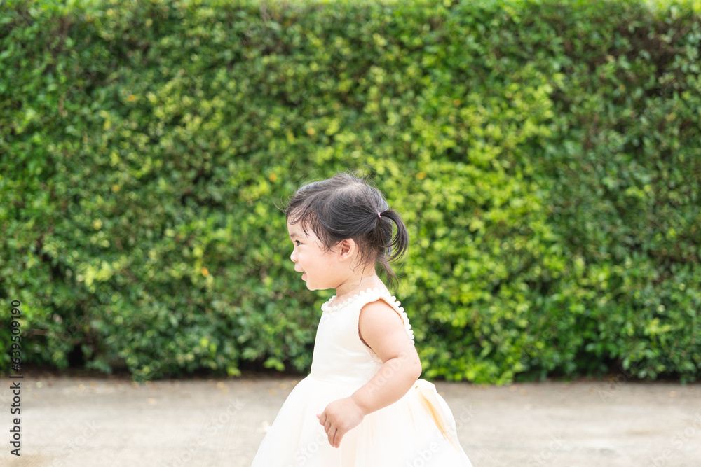 A cute little girl in beautiful white dress for wedding event in happy action on green leaf garden wall background. Kid portrait photo, face focus. A cute little girl in beautiful white dress for wedding event in happy action on green leaf garden wall background. Kid portrait photo, face focus.