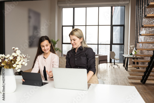 Portrait of mother and daughter sitting in living room. Cute girl holding in her hand a digital tablet while her mom using laptop and looking at something together.