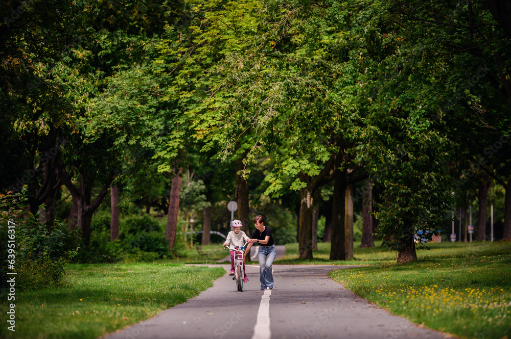Young mother in jeans and black t-shirt teaching and playing with her daughter in summer parks while girl riding a bike