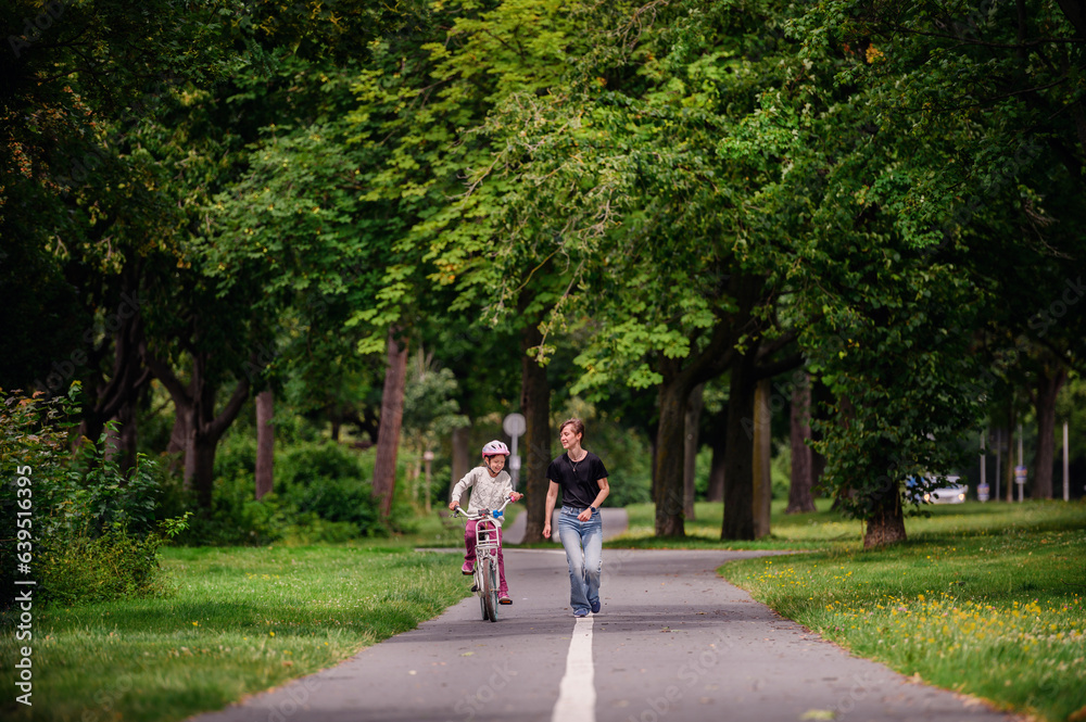 Young mother in jeans and black t-shirt teaching and playing with her daughter in summer parks while girl riding a bike