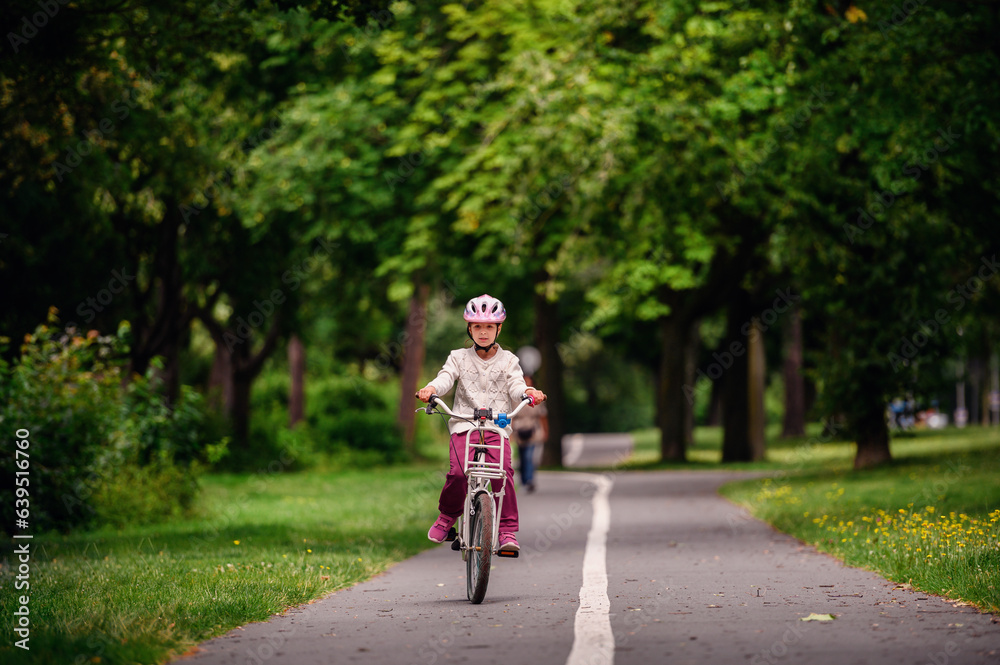 Little schooler girl riding bike in parks. Summer time, wearing helmet. From the back.