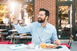 Handsome guy sitting in the restaurant and having a breakfast. Smiling man is holding a digital tablet and taking a selfie outdoors.