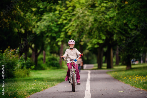 Little schooler girl riding bike in parks. Summer time, wearing helmet. From the back.