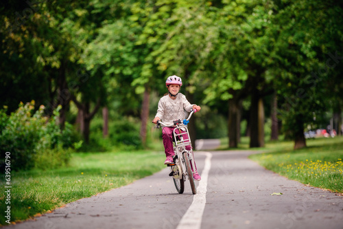 Little schooler girl riding bike in parks. Summer time, wearing helmet. From the back.