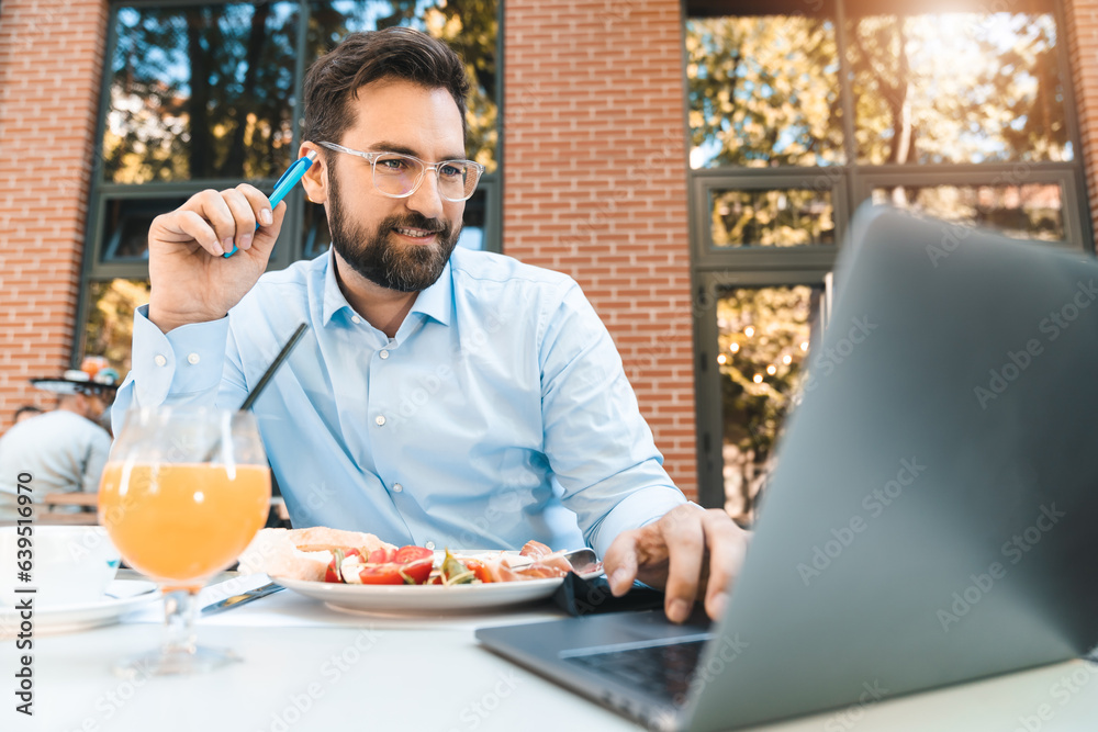 Focused businessman is using laptop and holding a pen. He is sitting in the restaurant outdoors and having a lunch break on a sunny day. Focused businessman is using laptop and holding a pen. He is sitting in the restaurant outdoors and having a lunch break on a sunny day.