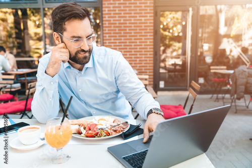 Smiling businessman is using laptop. He is sitting in the restaurant outdoors and having a lunch break on a sunny day.
