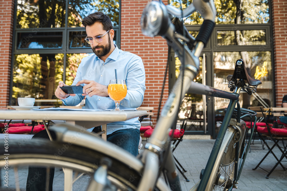 Obraz premium Businessman sitting and working in a local restaurant. He is sitting alone with a parked bicycle outside, drinking an orange juice and taking some photos with his phone.