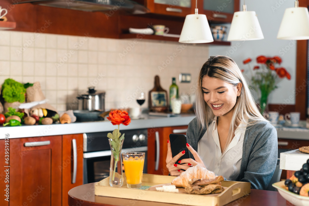Young smiling business woman using phone while having breakfast at home. Smiling female texting her partner or colleague during the lunch break.