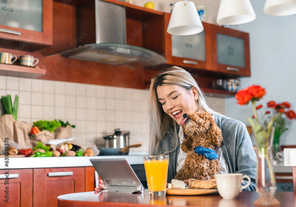 Young happy woman sitting at table and using tablet while holding a dog in her lap in kitchen. Groceries are visible on worktop in the back.
