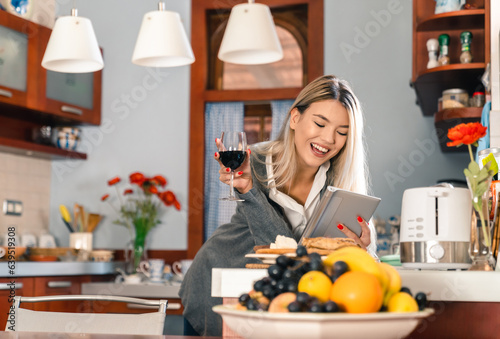 Satisfied young woman using digital tablet while drinking red wine in kitchen. Smiling girl spending her free time enjoying reading online magazine.