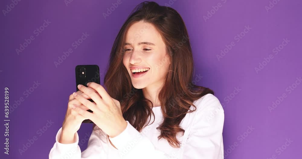 Smiling woman posing isolated on violet background in studio. Look coquettish hold using mobile cell phone. Girl flirting while chatting on mobile phone, dreaming looks up at the camera. Smiling woman posing isolated on violet background in studio. Look coquettish hold using mobile cell phone. Girl flirting while chatting on mobile phone, dreaming looks up at the camera.