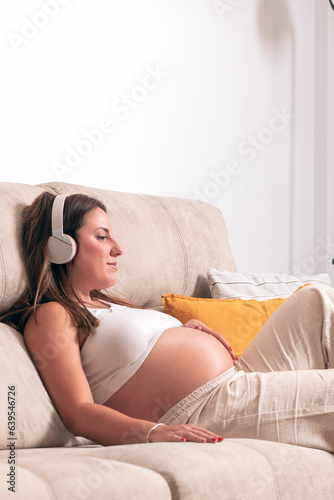 Young pregnant woman listening to music with headphones while relaxing on the couch at home