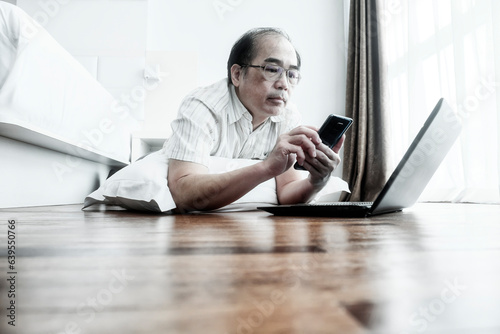 An adult man using his smartphone and laptop to work from home in his studio apartment, comfortably lying on the wooden floor.