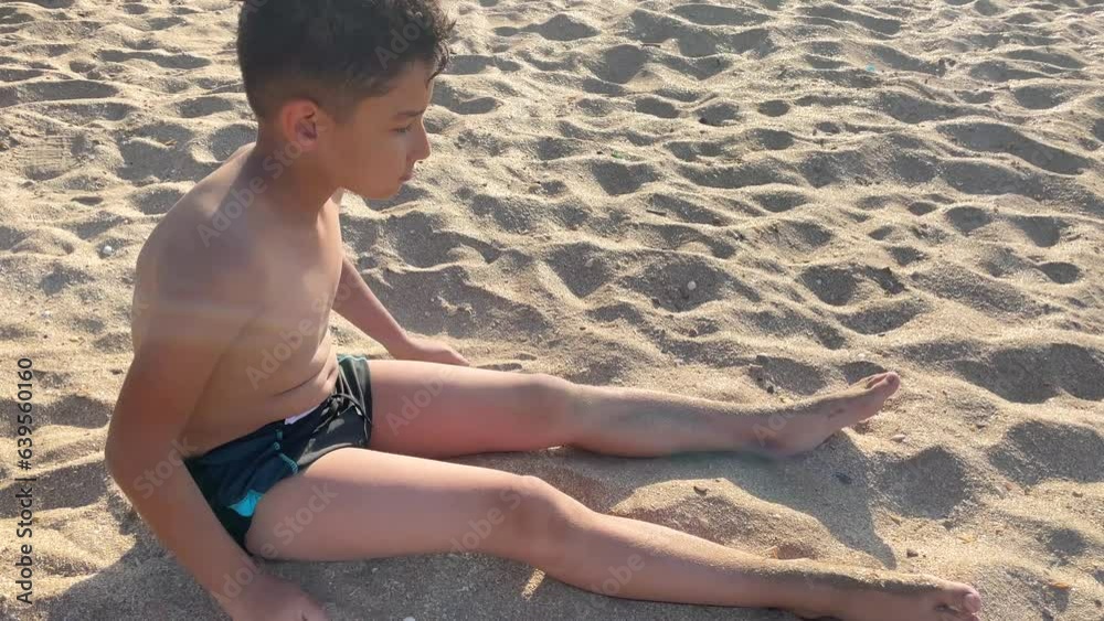 a 9-year-old boy on the beach. He sits on a sandy beach and sprinkles himself with sand