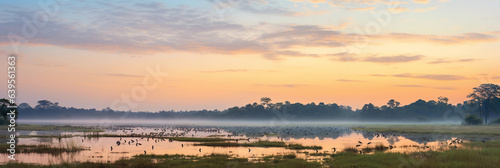 Serene panoramic view of a wildlife reserve at sunrise, showing a variety of animals grazing, soft pastel sky
