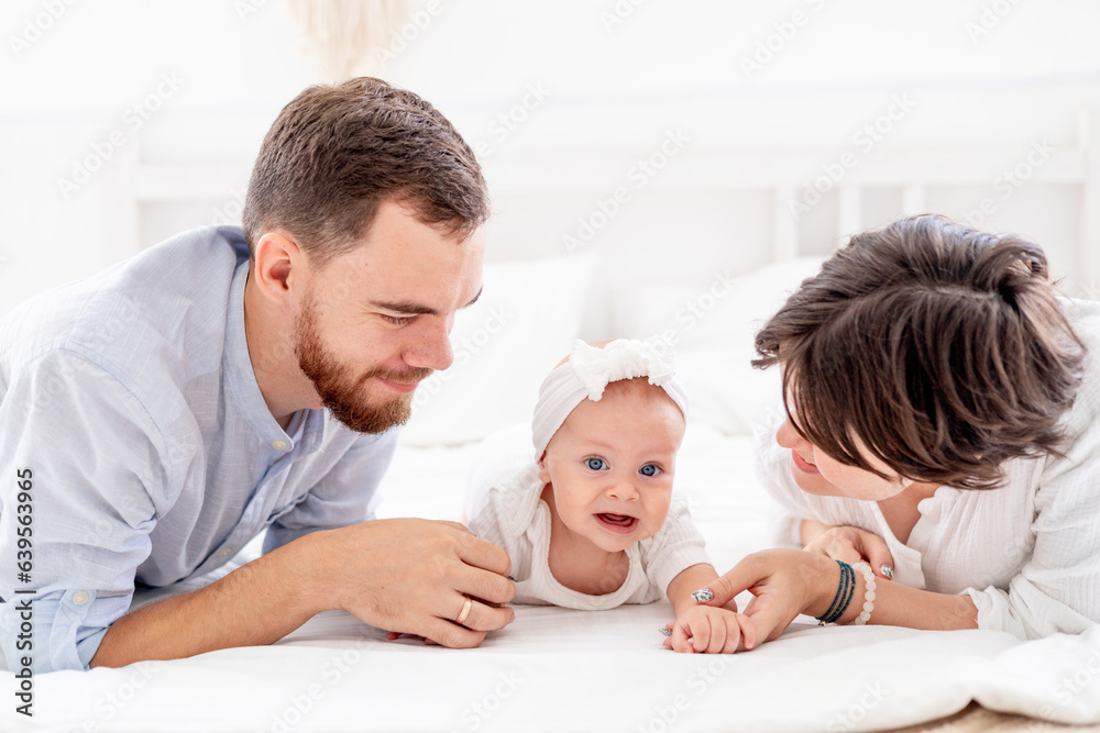 a happy young family with a newborn baby hugging and kissing on the bed at home in a bright room, mom, dad and a small child on a white bed smiling a happy young family with a newborn baby hugging and kissing on the bed at home in a bright room, mom, dad and a small child on a white bed smiling