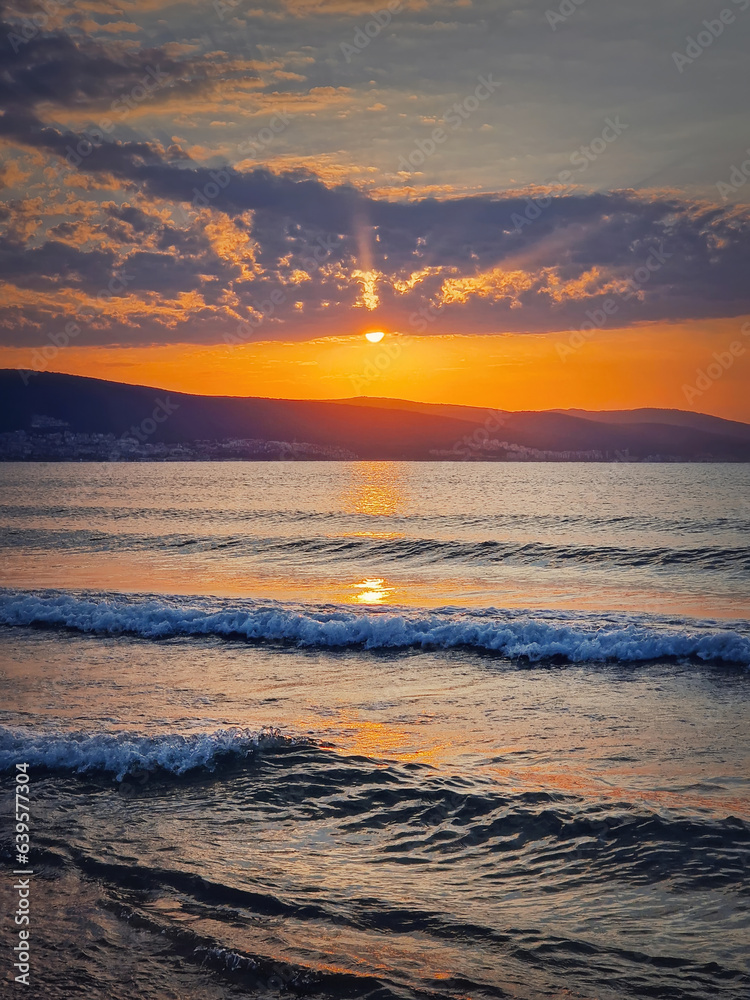 Fototapeta premium Vibrant sunrise at the Bulgarian coastline of Black Sea. Sunny beach resort, dawn scene with the sun rising up the hills on the horizon and foamy waves washing the sand shore