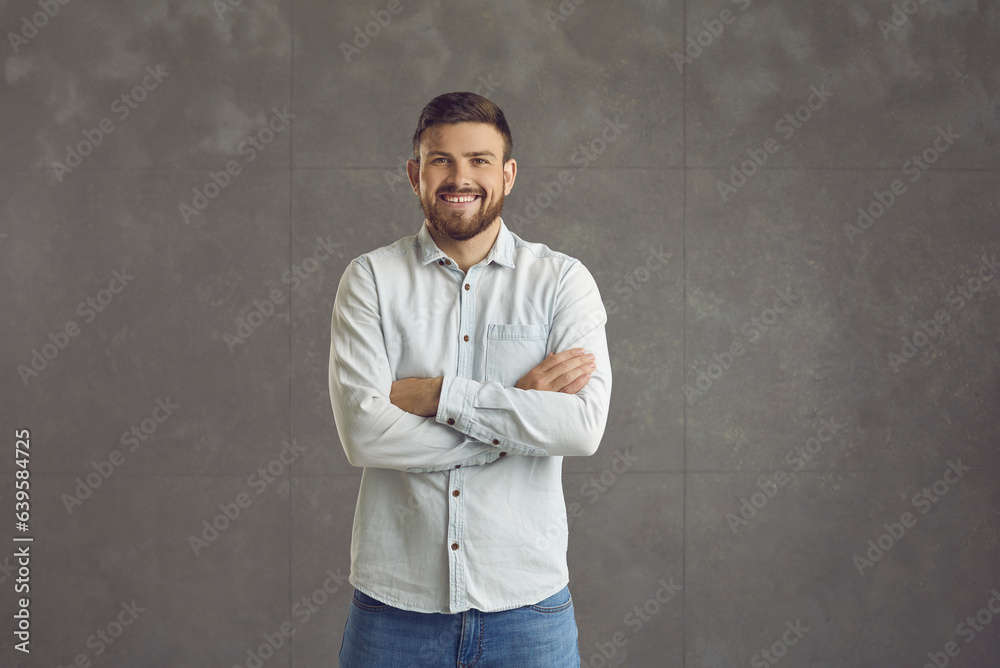 Portrait of happy charming young man in casual denim outfit standing arms crossed and smiling at camera. Studio shot of handsome bearded guy in his 20s against background of grey studio wall
