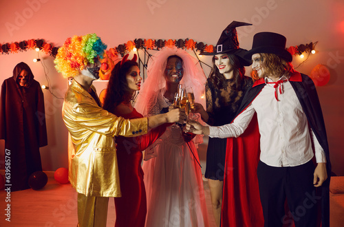 Group of cheerful young people together clinking glasses of champagne at party for Halloween. Multiracial friends dressed in spooky costumes have fun together at home. Halloween celebration concept.