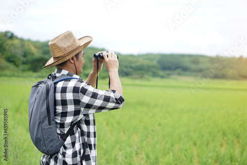 Asian man naturalist wears hat, plaid shirt, backpack, uses binocular to explore nature at paddy field. Concept, nature exploration. Ecology study. Pastime activity, lifestyle. Explore environment. 