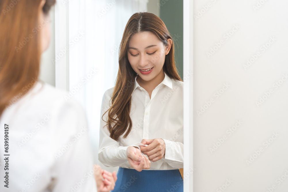 Get dress, asian young woman, businesswoman standing, hand buttoning up shirt formal, female getting dressed, ready before go to work looking reflection the mirror in bedroom in the morning at home. Get dress, asian young woman, businesswoman standing, hand buttoning up shirt formal, female getting dressed, ready before go to work looking reflection the mirror in bedroom in the morning at home.