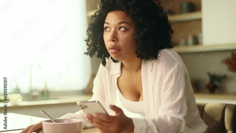 Boring exhausted curly woman watching uninteresting online videos programs or news in social media on smartphone gadget while having breakfast in morning at home Tired female after bad sleep insomnia Boring exhausted curly woman watching uninteresting online videos programs or news in social media on smartphone gadget while having breakfast in morning at home Tired female after bad sleep insomnia