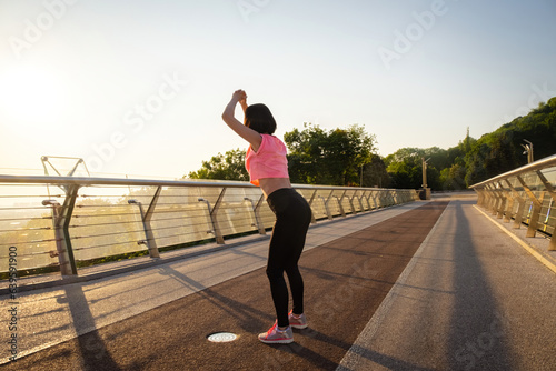 Young sportswoman takes hands above head and starts squat exercise. Morning gymnastics at sunrise on footbridge in summer