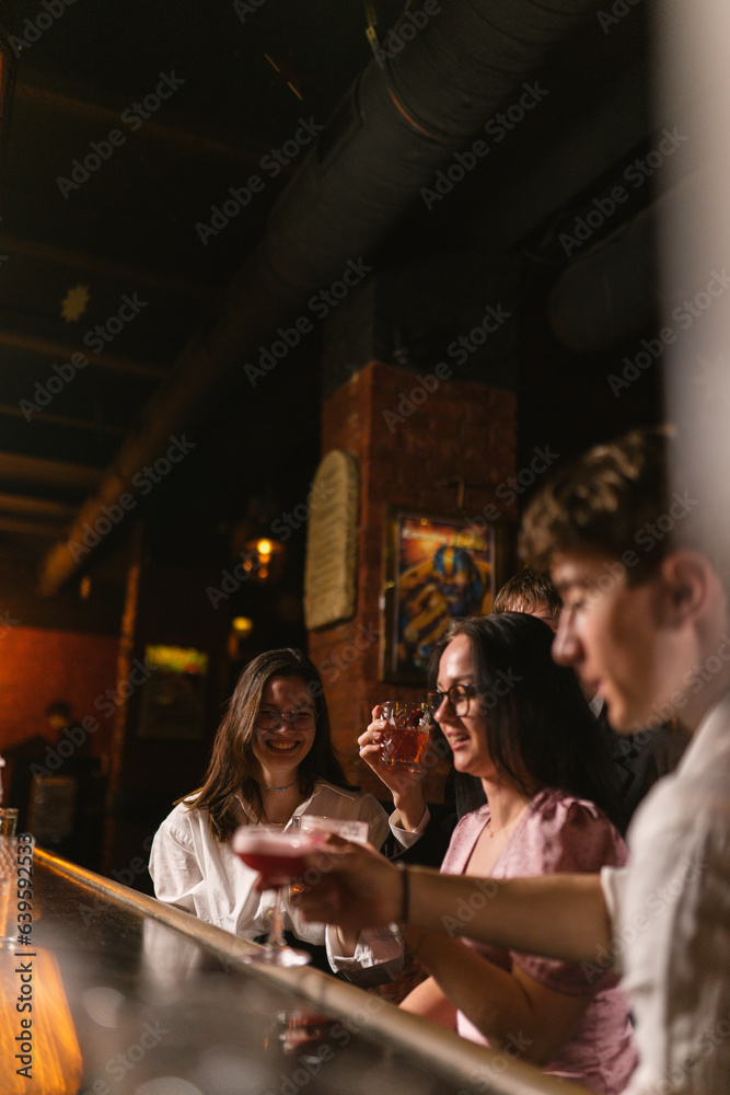 Guys and girls celebrate meeting in fancy restaurant. Young people drink. Girls tell funny jokes to friends at bar counter