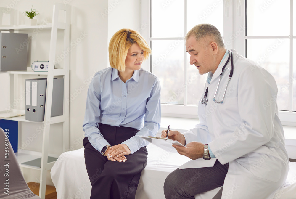 Obraz premium Doctor talking to patient. Senior physician in white coat sitting together with young woman on medical bed in examination room, holding clipboard and talking about health check up results