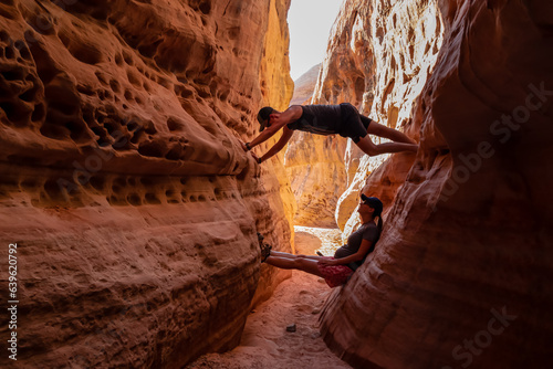 Couple climbing the walls in narrow Kaolin Wash slot canyon on White Domes Hiking Trail in Valley of Fire State Park in Mojave desert, Nevada, USA. Massive cliffs of striated red white rock formations