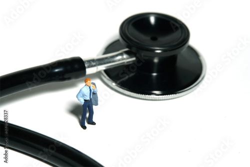 Miniature tiny people toy figure photography. A men standing in front of stethoscope, waiting for doctor health care check. Isolated on white background