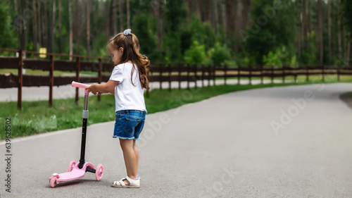 Seasonal children's sports. Healthy childhood lifestyle. Summer holidays. Little girl, child riding a scooter on the road in the park outdoors.