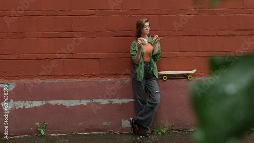 Skater girl resting enjoying street food. Young woman eating burger standing near urban wall with skateboard during rain. Wide angle shot.  photo