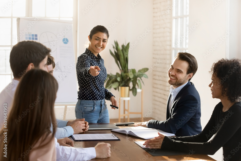 Obraz premium Smiling female indian team leader interact with employee at group briefing in office, happy excited diverse multiracial colleagues participate in teambuilding activity at meeting or training together