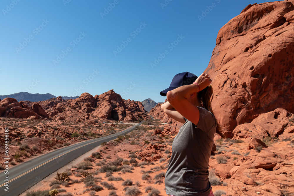 Woman standing on endless winding empty Mouse tank road in Valley of Fire State Park through canyons of red Aztec Sandstone Rock formations and desert vegetation in Mojave desert, Overton, Nevada, USA