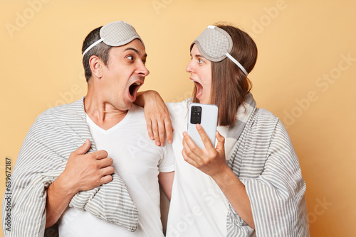 Shocked surprised couple man and woman in sleep eye mask wrapped in blanket isolated over beige background using cell phone after waking up browsing internet screamign with amazement. photo