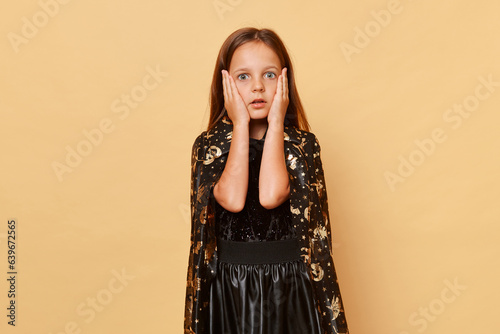 Canvas Print Shocked scared little girl wearing witch costume isolated over beige background keeps hands on cheeks looking with big eyes at something scary halloween celebration