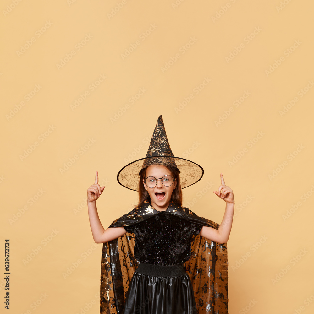 Excited crazy little girl wearing witch costume carnival cone hat and glasses isolated over beige background screaming with amazement pointing up at advertisement area.