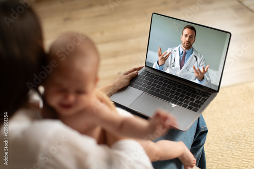 Caring mother holding crying baby and using laptop, making video call to pediatrician at home, doctor consulting online. Telemedicine and child healthcare concept
