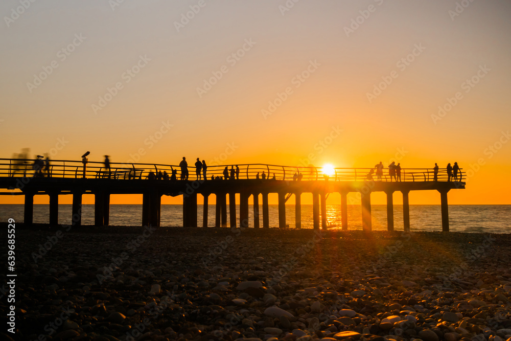 Silhouettes of people are walking on the pier against the clear sky over the Black Sea at sunset. Summer, peaceful, vacation and sightseeing concept