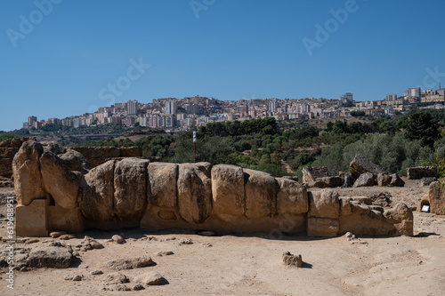 Hellenic statue of a rocky giant  atlas  lying on the sand at the valley of Temples in Agrigento  Sicily. City skyline in the background 