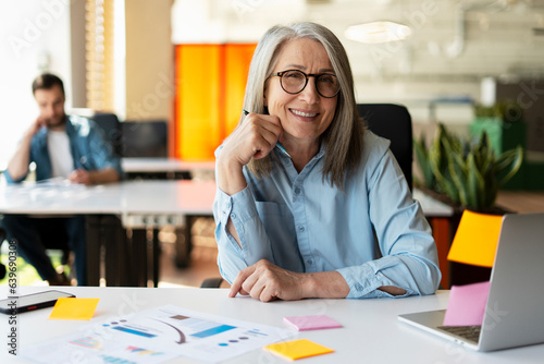 Portrait of smiling confident senior businesswoman using sticky notes looking at camera sitting in modern office. Gray haired manager wearing stylish eyeglasses  at workplace. Scrum, agile concept