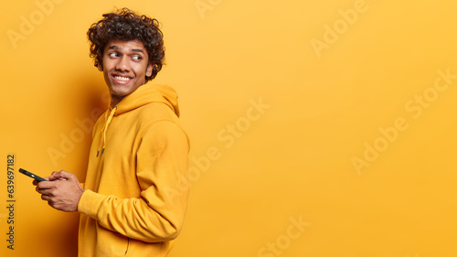 Cheerful Hindu man using cellphone and smiling reading good news message enjoying mobile application looks behind dressed in casual sweatshirt isolated over yellow background copy space for your promo