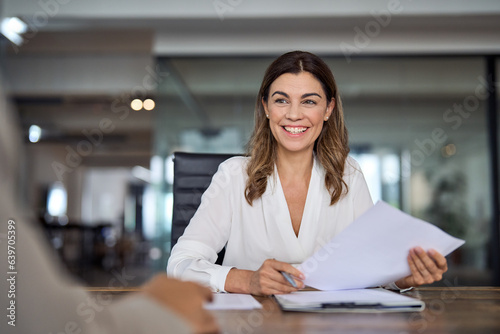 Smiling mature business woman hr holding cv document at job interview. Happy mid aged professional banking manager or lawyer consulting client sitting at workplace in corporate office meeting.