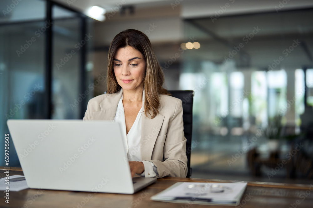 © insta_photos - Busy mature middle aged professional business woman manager executive looking at laptop computer technology in office working on digital financial banking market sitting at desk. Copy space.